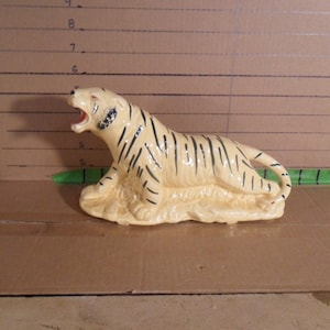 May include: A ceramic tiger figurine in a reclining pose, with an open mouth. The tiger has a cream-colored body with black stripes and is resting on a cream-colored base. The figurine is a decorative item.