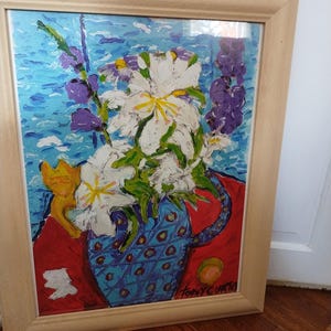 May include: An original framed painting of white flowers in a blue vase with a cat figurine. The painting has a light wood frame and is set against a blue background. The vase is blue with a diamond pattern.