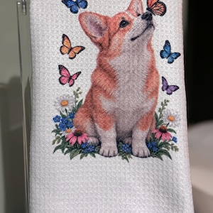 May include: A white waffle-weave kitchen towel featuring a printed illustration of a tan and white Corgi sitting among colorful wildflowers. The dog looks upward at a monarch butterfly, surrounded by additional blue, orange, and purple butterflies.