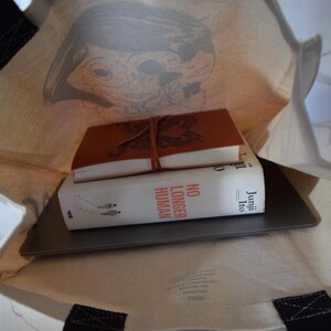 May include: A white canvas tote bag with black handles. The bag is filled with a laptop, a book titled "No Longer Human" by Junji Ito, and a brown leather-bound journal.