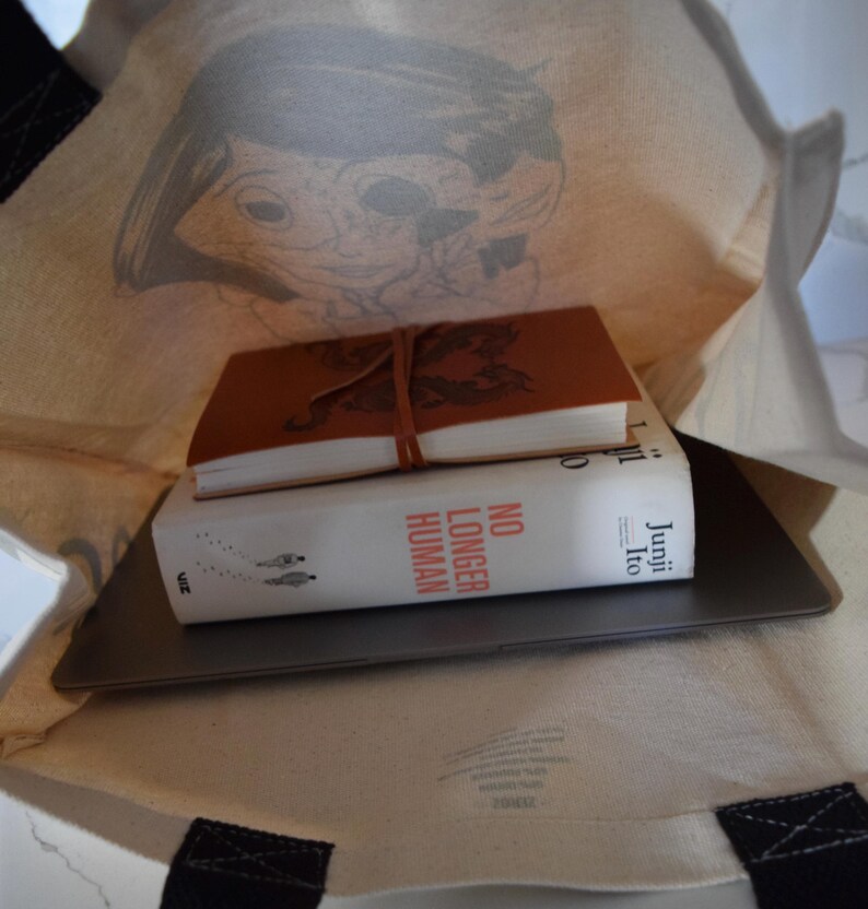 May include: A white canvas tote bag with black handles. The bag is filled with a laptop, a book titled "No Longer Human" by Junji Ito, and a brown leather-bound journal.