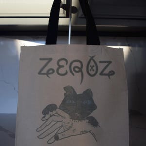 May include: A white canvas tote bag with black handles. The bag has a black cat with blue eyes printed on it. The text "ZEROZ" is printed in black above the cat.