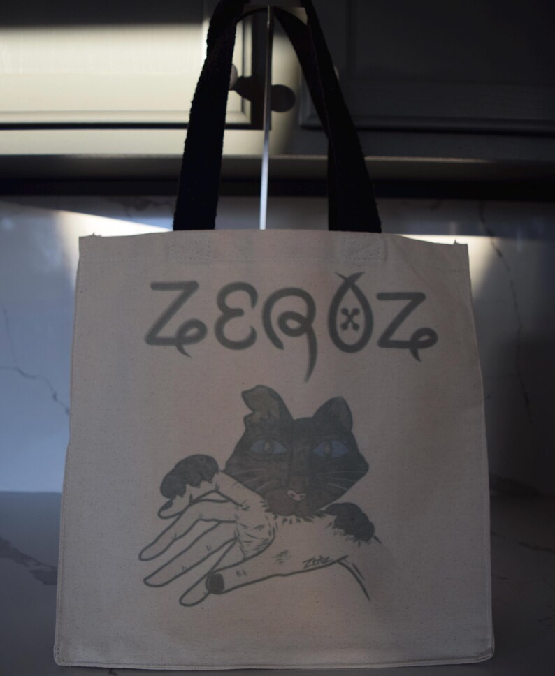May include: A white canvas tote bag with black handles. The bag has a black cat with blue eyes printed on it. The text "ZEROZ" is printed in black above the cat.