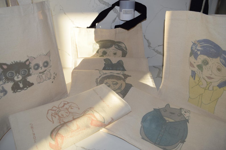 May include: A set of six tote bags with various cartoon illustrations printed on them. The illustrations include a cat, a crab, a monster, a girl with multiple faces, and a blue creature. The bags are made of natural canvas fabric.