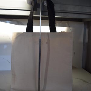 May include: A plain white canvas tote bag with black fabric handles. The bag is hanging on a metal stand.