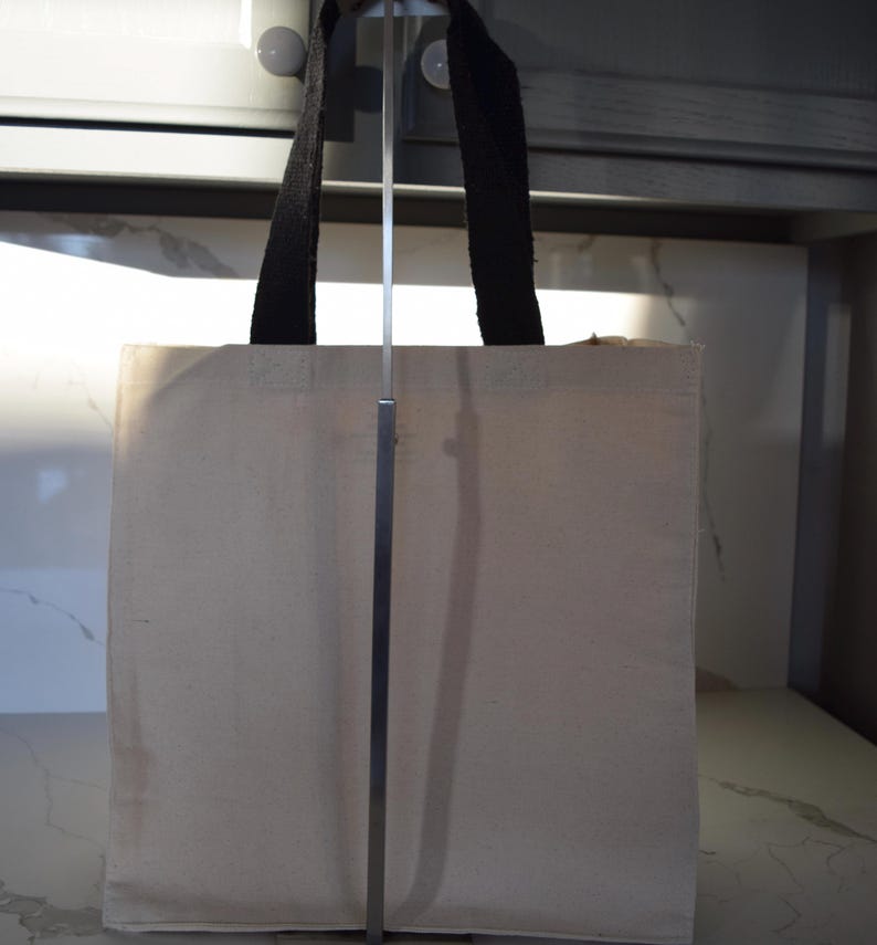 May include: A plain white canvas tote bag with black fabric handles. The bag is hanging on a metal stand.