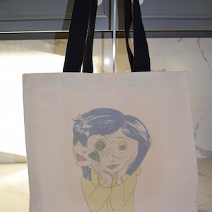 May include: A white canvas tote bag with black straps featuring a colorful illustration of a girl with blue hair and a yellow raincoat. The girl's face is partially obscured by a large eye.