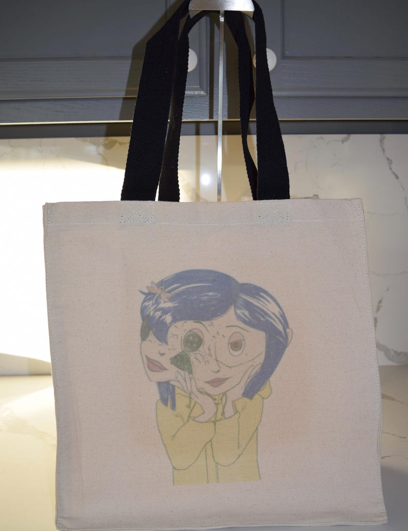 May include: A white canvas tote bag with black straps featuring a colorful illustration of a girl with blue hair and a yellow raincoat. The girl's face is partially obscured by a large eye.