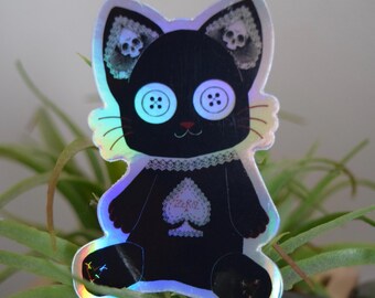 Puede incluir: Pegatina de gato negro con ojos de botón, cuello de encaje y diseño de pica. Las orejas del gato presentan diseños de calaveras. La pegatina tiene un acabado holográfico iridiscente, sobre un fondo de plantas verdes.