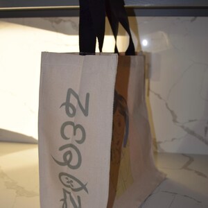 May include: A beige canvas tote bag with black handles. The bag has a green design on the front that says "Zegna 2023".