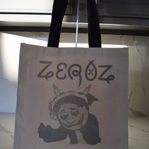 May include: A white canvas tote bag with black handles. The bag has a cartoon character with a cigarette and candles on its head. The text "ZEEQOZ" is printed on the bag.