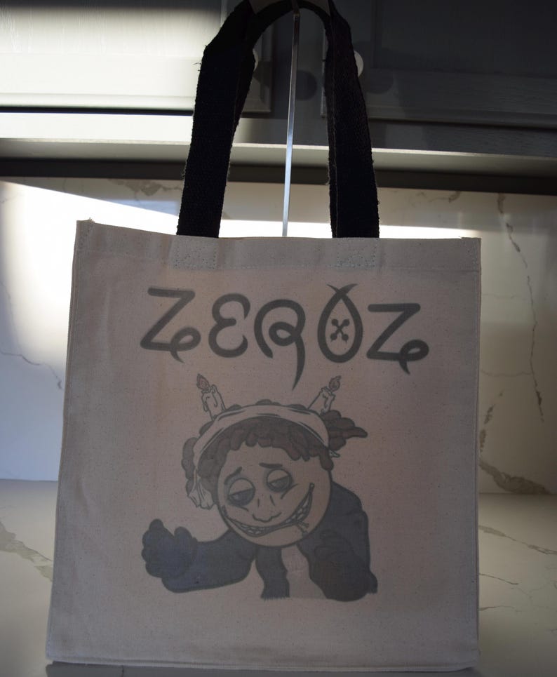 May include: A white canvas tote bag with black handles. The bag has a cartoon character with a cigarette and candles on its head. The text "ZEEQOZ" is printed on the bag.