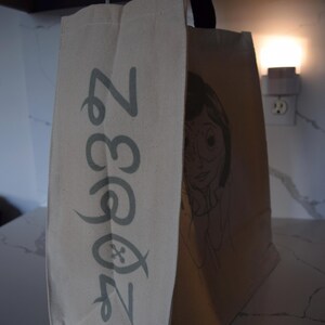 May include: A beige canvas tote bag with a black handle. The bag has a green and white logo that says "Zoo 33" and a black and white illustration of a girl with glasses on the side.