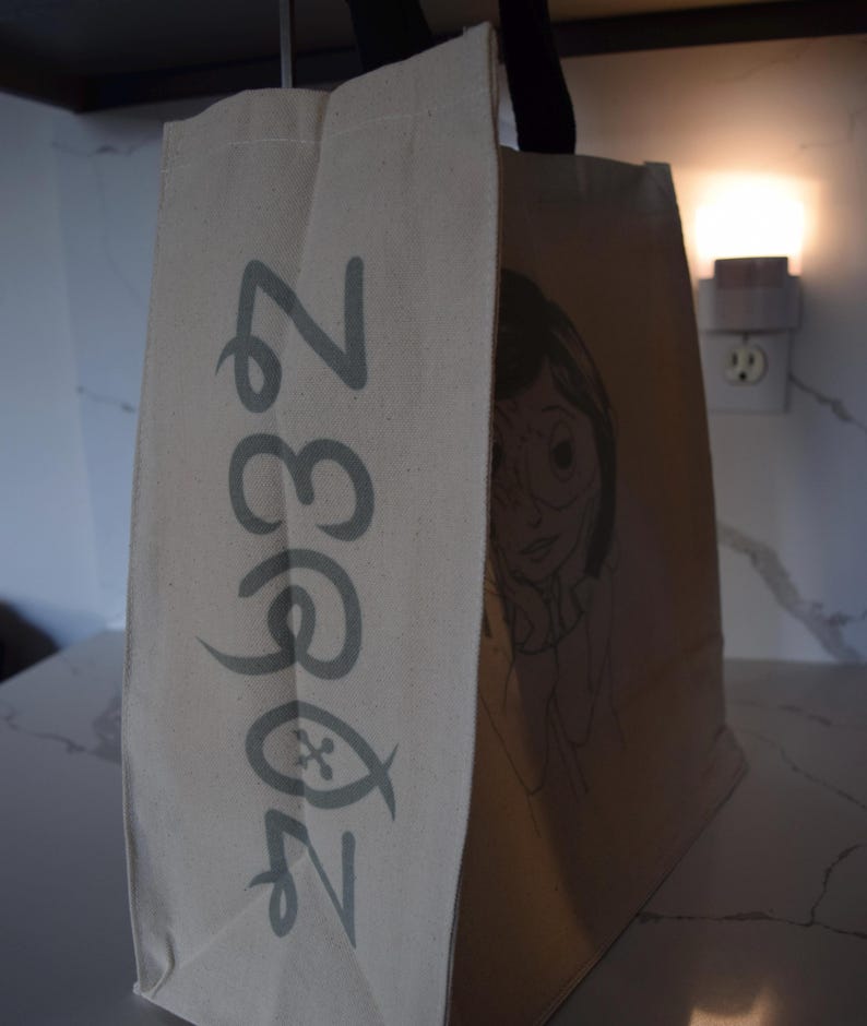 May include: A beige canvas tote bag with a black handle. The bag has a green and white logo that says "Zoo 33" and a black and white illustration of a girl with glasses on the side.