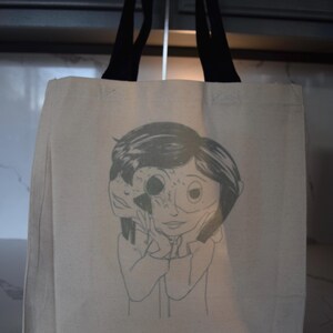 May include: A beige canvas tote bag with black handles. The bag features a black and white illustration of a girl with a face that is partially obscured by a mask.