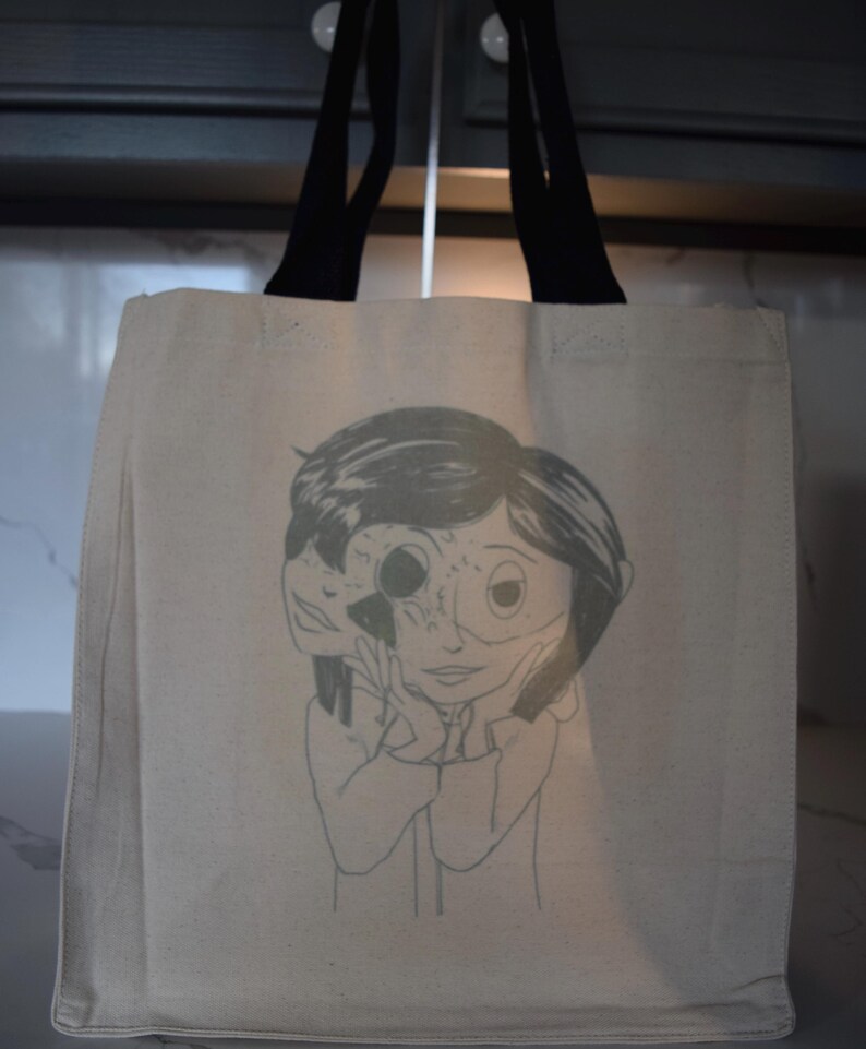 May include: A beige canvas tote bag with black handles. The bag features a black and white illustration of a girl with a face that is partially obscured by a mask.