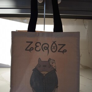 May include: A beige canvas tote bag with black handles. The bag has a cartoon illustration of a gray cat wearing a blue shirt and a hockey mask. The text "ZEROZ" is printed on the bag.