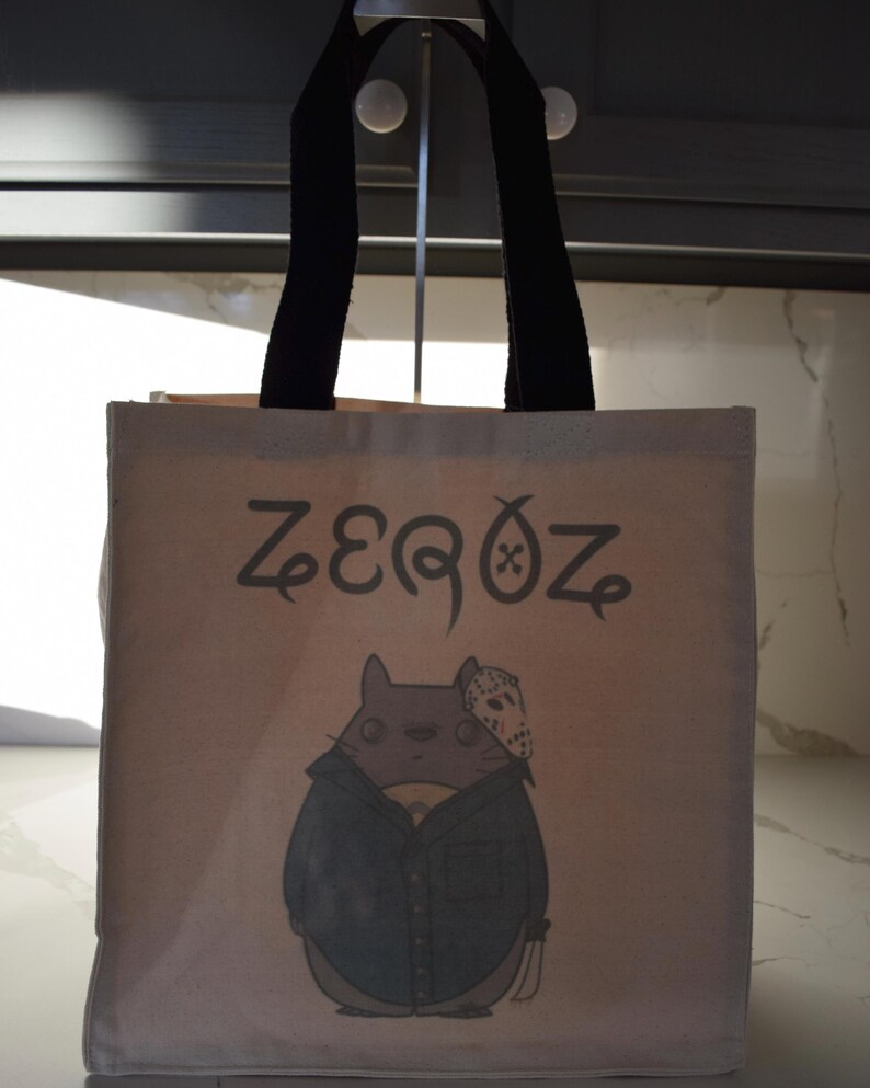 May include: A beige canvas tote bag with black handles. The bag has a cartoon illustration of a gray cat wearing a blue shirt and a hockey mask. The text "ZEROZ" is printed on the bag.