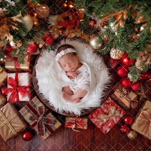 Puede incluir: Un bebé recién nacido dormido, envuelto en encaje blanco y con una diadema floral, en un nido blanco y esponjoso. Rodeado de decoraciones navideñas, incluyendo regalos envueltos, adornos y un árbol de Navidad.