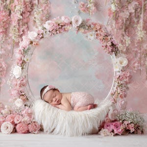 May include: A newborn baby sleeps peacefully on a fluffy white surface inside a floral arch. The arch is adorned with pink and white flowers, set against a soft pink and blue backdrop. The baby wears a pink headband and matching diaper cover.