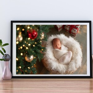 May include: A framed photograph featuring a newborn baby wrapped in a white blanket, nestled in a fluffy, round basket. A Christmas tree with ornaments and lights is in the background, along with wrapped gifts.