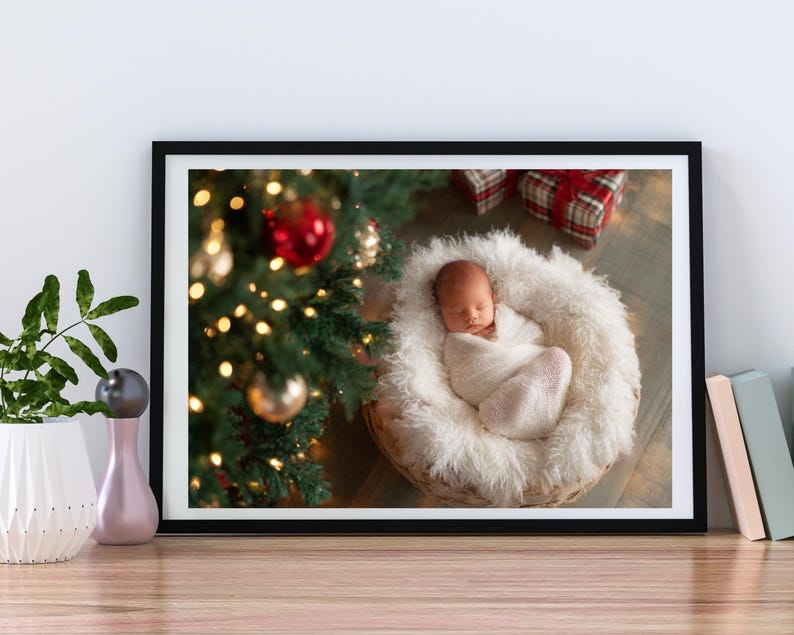 May include: A framed photograph featuring a newborn baby wrapped in a white blanket, nestled in a fluffy, round basket. A Christmas tree with ornaments and lights is in the background, along with wrapped gifts.