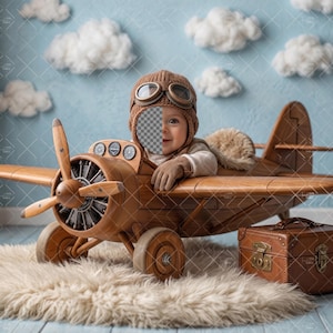 May include: A wooden toy airplane with a propeller, wheels, and a small brown suitcase. The plane has a pilot's seat and is set against a blue backdrop with fluffy white clouds. A child is wearing a brown aviator hat and gloves.
