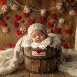Op de afbeelding: Een slapende baby die een witte gebreide muts en een witte gebreide deken draagt, ligt in een houten vat met een bordje waarop "My Valentine" staat. Het vat is omringd door rode vilt hartjes.