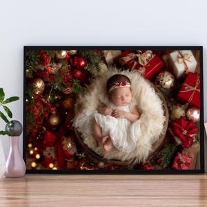 May include: A framed photograph of a newborn baby in a white dress and headband, nestled in a fluffy basket. The baby is surrounded by Christmas decorations, including red and gold ornaments and wrapped gifts. The frame is black.