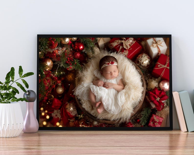 May include: A framed photograph of a newborn baby in a white dress and headband, nestled in a fluffy basket. The baby is surrounded by Christmas decorations, including red and gold ornaments and wrapped gifts. The frame is black.