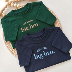 Peut inclure: Un t-shirt vert foncé et un t-shirt bleu marine, tous deux à manches courtes et à col rond. Chaque t-shirt présente le texte brodé "est. 2027" au-dessus des mots "big bro." dans une couleur de fil assortie au design de chaque t-shirt.