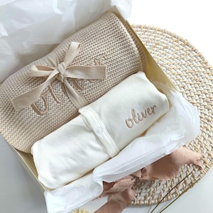 Könnte beinhalten: Ein beiges Baby-Geschenkset mit Decke und Strampler. Die Decke ist mit einem Band gebunden und mit dem Namen "Olivia" bestickt. Der weiße Strampler ist ebenfalls mit dem Namen "Oliver" bestickt. Die Box ist mit Seidenpapier ausgekleidet.