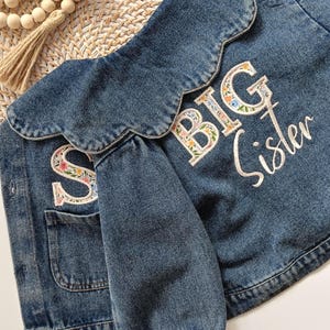 Puede incluir: Una chaqueta de mezclilla azul con cuello festoneado y mangas largas con puños elásticos. La parte posterior de la chaqueta está bordada con el texto BIG Sister en hilo blanco, con las letras rellenas de un patrón floral colorido sobre un fondo blanco.