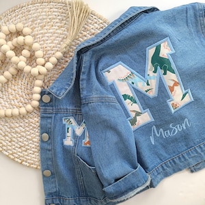 May include: Pale blue denim jacket with a large letter "M" patch on the back and a smaller one on the pocket. The patches feature a dinosaur pattern. The name "Mason" is embroidered in cursive below the large letter.