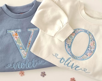 May include: A blue sweatshirt with an embroidered letter V and the name Violet, and a cream sweatshirt with an embroidered letter O and the name Olive. Both feature floral patterned fabric within the letters and light blue cursive embroidery.