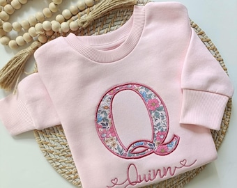 May include: A light pink long-sleeve sweatshirt featuring a large embroidered letter Q with a floral pattern in shades of pink, blue, and green. Below the letter, the name Quinn is embroidered in cursive script with small heart accents.