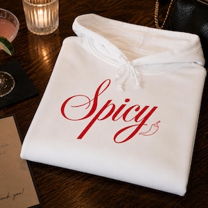 Op de afbeelding: Een witte hoodie met het woord "Spicy" in rode letters en een rode chilipeper. De hoodie ligt op een donker houten oppervlak, naast een cocktail en een menu.