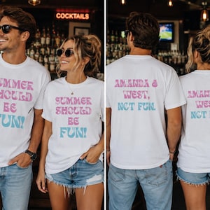 以下が含まれることがあります： 白いTシャツを着た2人。Tシャツの前面にはピンクと青で「SUMMER SHOULD BE FUN！」と書かれています。Tシャツの背面には「AMANDA & WEST、NOT FUN」と書かれています。背景はバーのようです。