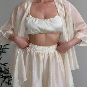 Op de afbeelding: Een cr&egrave;mekleurige driedelige pyjamaset. De set bevat een top in camisole-stijl, een short en een lange, openvallende kimono. De stof lijkt een lichtgewicht, transparant materiaal te zijn, met een subtiele glans. De short heeft een elastische tailleband.