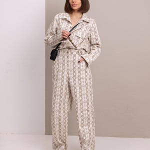 Könnte beinhalten: Ein weißer und brauner Jumpsuit mit Schlangenmuster, einem Kragen und langen Ärmeln. Der Jumpsuit hat einen Knopfverschluss und Taschen. Das Model trägt schwarze Sandalen.