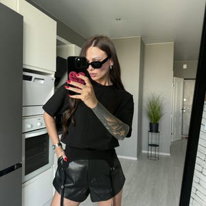 May include: A person wearing a black t-shirt, black leather shorts with suspenders, and black sunglasses. The person is holding a phone with a pink heart design. The person is wearing black stockings.