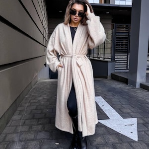 Op de afbeelding: Een lange, beige cardigan met bijpassende riem, gedragen over een zwarte top en legging. De cardigan heeft lange mouwen en een relaxte pasvorm. De persoon draagt zwarte laarzen en een zonnebril. De outfit is geschikt voor herfst of winter.