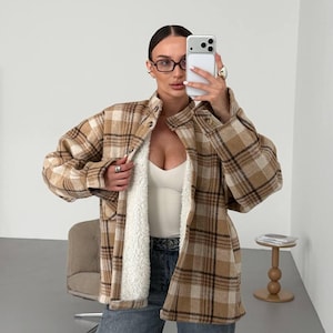 May include: A tan and brown plaid button-down shirt jacket featuring a white sherpa fleece lining, a fold-over collar, long sleeves with buttoned cuffs, and two chest pockets. The garment is worn over a white tank top and blue denim jeans.