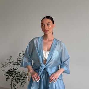 Op de afbeelding: Een lichtblauwe, semi-transparante driedelige outfit. De set bevat een kimono-top met strik, een bijpassende short en een witte crop top. De outfit is gemaakt van een lichte stof en is geschikt voor warm weer.