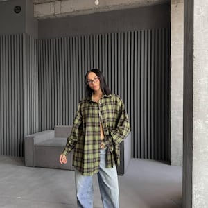Könnte beinhalten: Eine Person trägt ein grün-schwarzes kariertes Hemd mit langen Ärmeln, blaue Jeans und schwarz-weiße Sneaker. Das Hemd ist überdimensioniert und hat eine lockere Passform.