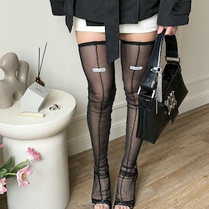 Puede incluir: Medias altas negras transparentes con costuras verticales y el texto "The Creator" en la liga. La persona lleva sandalias negras de tacón alto y sostiene un bolso negro con herrajes plateados.