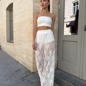 May include: A white two-piece outfit consisting of a strapless top and wide-leg lace pants. The pants feature a floral lace pattern. The outfit is worn with white sandals. The background is a beige building with a door.