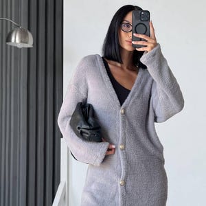 Può includere: Un cardigan grigio chiaro a maniche lunghe con bottoni dorati. Il cardigan è realizzato in un materiale morbido e soffice. Una pochette nera è tenuta nella mano sinistra. La persona indossa leggings neri e un top nero.