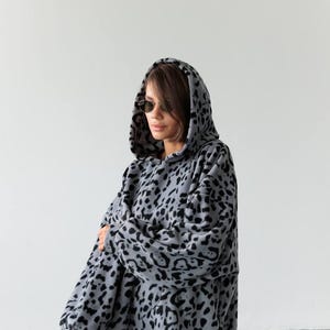 Pode incluir: Moletom cinza com estampa de leopardo preto. O moletom oversized tem capuz grande e mangas compridas. A modelo usa óculos de sol e leggings pretas.