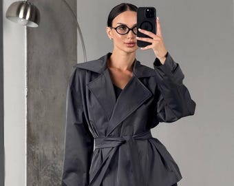 May include: A dark gray jacket with a wrap-around belt. The jacket has a wide collar and long sleeves. The person in the image is wearing black leggings and glasses, holding a black phone.